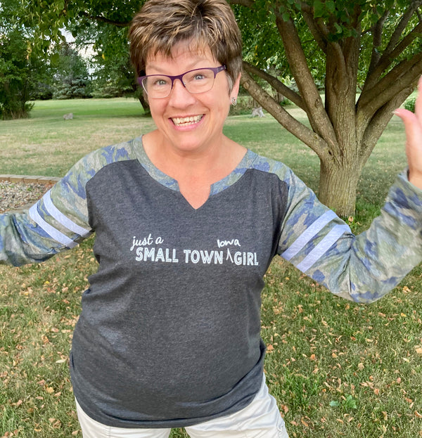 A ladies cut long sleeve notched collar jersey styled tee shirt. Dark grey body with Camo sleeves . Front white graphic says Just A Small Town Iowa Girl .