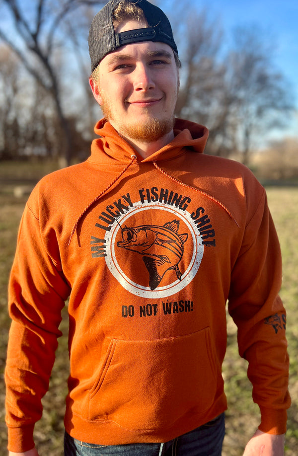 A man wearing a Texas Orange colored, hood, pouch front, unisex sized sweatshirt. The Large front graphic, done in light grey and black is of a large, active fish with a hook in its mouth. Wording, in an arc over and below the fish reads MY LUCKY FISHING SHIRT, DO NOT WASH! Available in sizes small to 3X-Large.