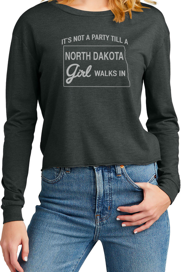 Woman wearing a long sleeved tee that is cropped below the belly button. Shirt color is Black Frost. Crew neck, cuffs on sleeves, surged hem at bottom of shirt. Front white graphic reads, It's Not A Party Till A North Dakota Girl Walks In. Cotton, polyester, rayon blend shirt. Sizes Extra small to 3 XL.