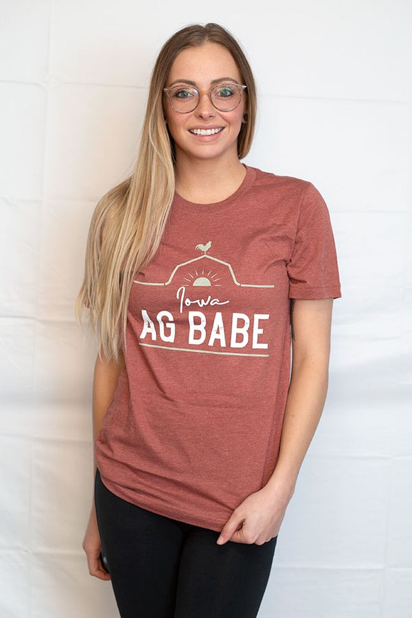 A short sleeved unisex crew neck tee shirt in Heather Clay color. The graphic shows a rooster perched on a barn roof at sunrise. The words Iowa Ag Babe are printed on the shirt front.