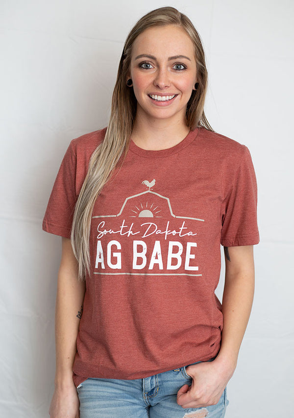 A short sleeved unisex crew neck tee shirt in Heather Clay color. The graphic shows a rooster perched on a barn roof at sunrise. The words South Dakota Ag Babe are printed on the shirt.