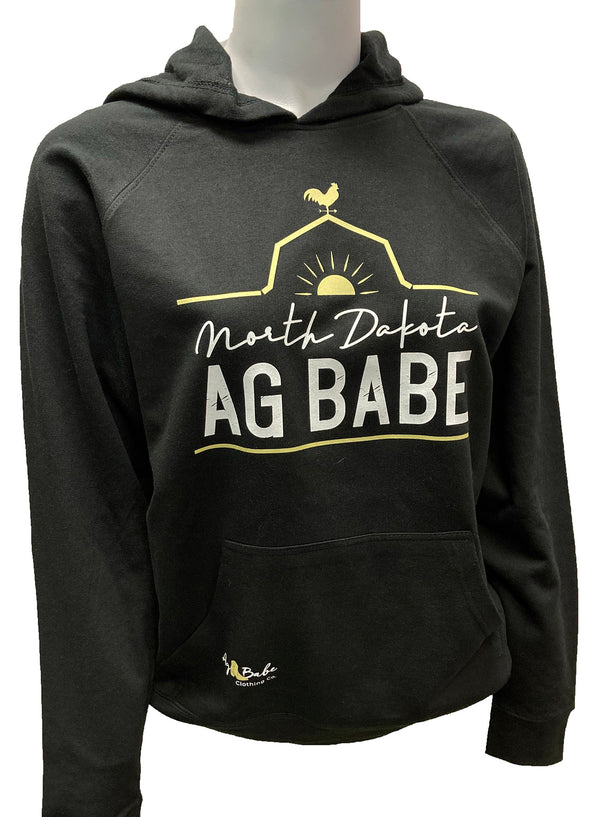 A mannequin wearing a unisex, black pouch front hooded sweatshirt. A front graphic in gold and white shows a barn roof line, rooster and sun with sun rays. Just under the roof line large lettering reads North Dakota Ag Babe  