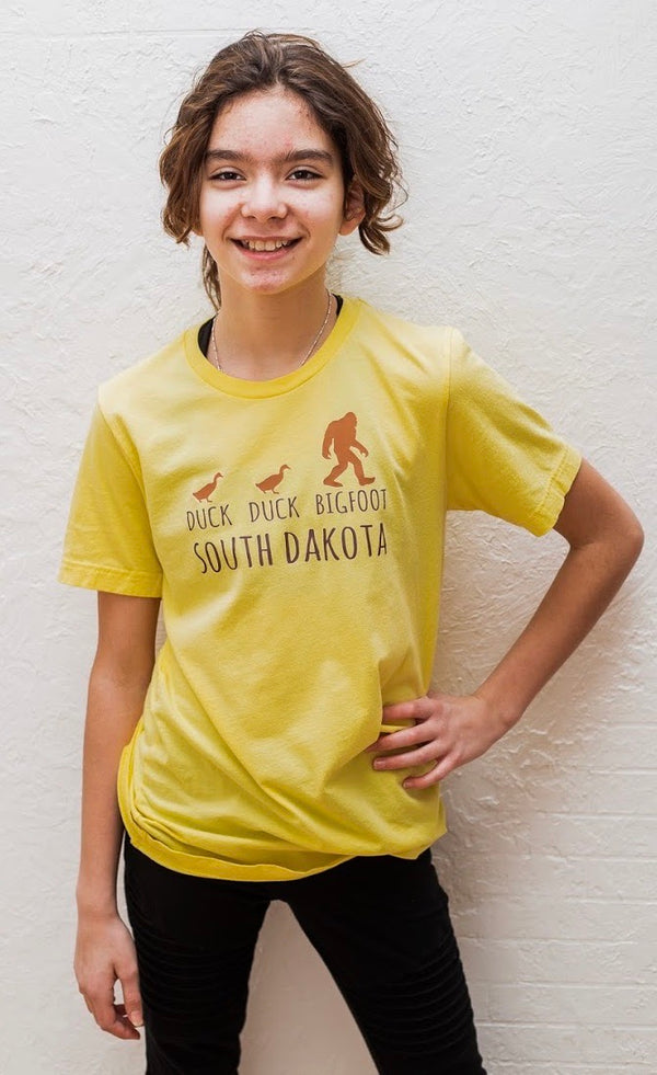 A Heather Yellow Gold short sleeved toddler and youth sized shirt. The front graphic shows two ducks following Bigfoot. The design is called Duck Duck Bigfoot South Dakota.