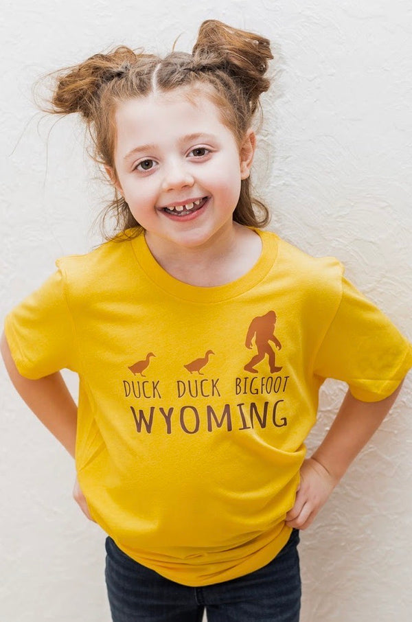 A Heather Yellow Gold short sleeved toddler and youth sized shirt. The front graphic shows two ducks following Bigfoot. The design is called Duck Duck Bigfoot Wyoming.