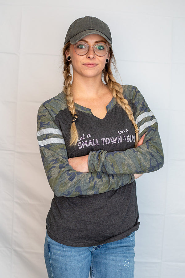 A young woman wearing a ladies cut long sleeve notched collar jersey styled tee shirt. Dark grey body with Camo sleeves . Front  white graphic says Just A Small Town Iowa Girl .
