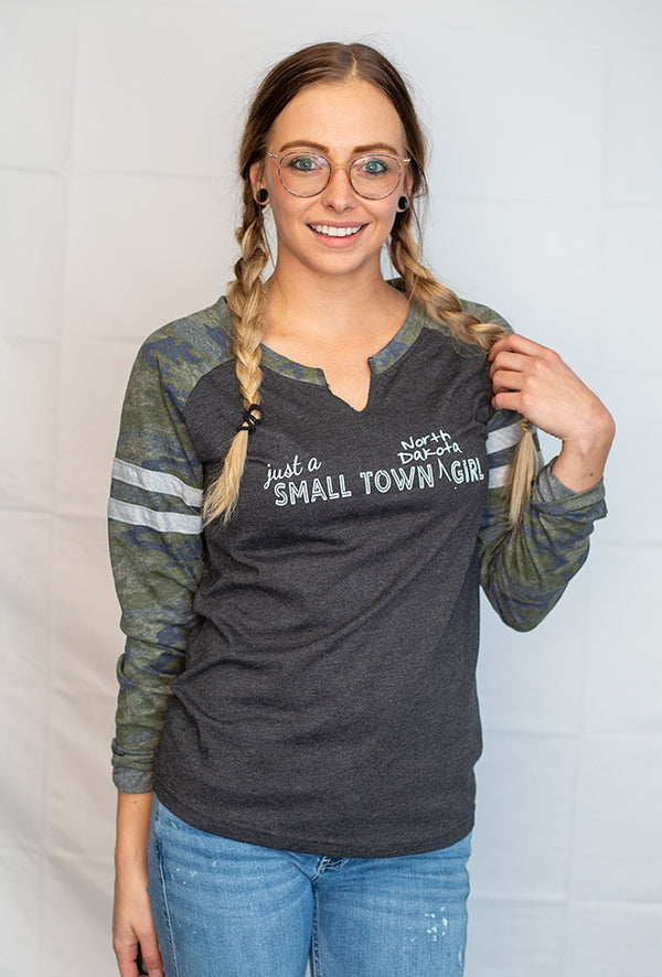 A ladies cut long sleeve notched collar jersey styled tee shirt. Dark grey body with Camo sleeves . Front white graphic reads Just A Small Town North Dakota Girl .