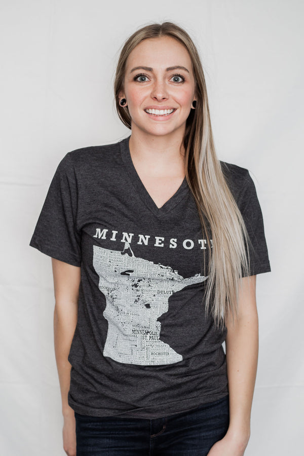 A woman wearing a unisex sized,Dark Grey Heather V neck, cotton polyester, short sleeve tee. Front white graphic is of Minnesota and its cities. Sizes small to 3X-Lagre. A Scratchpad Tees original design.