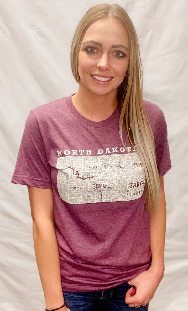 A woman wearing a unisex sized, Heather Maroon crew neck, cotton polyester, short sleeve tee. Front light grey graphic is of North Dakota and its cities. Sizes small to 3X-Lagre. A Scratchpad Tees original design.