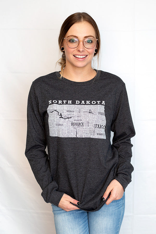 A woman wearing a unisex, long sleeved Dark Grey Heather, cotton polyester tee shirt. The front graphic, done in light grey is of the state of North Dakota and most of its cities. Scratchpad Tees original design available in sizes small to 3X-Large.
