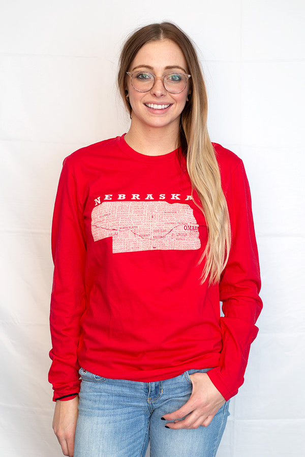 A woman wearing a 100 percent cotton, red, unisex long sleeved tee shirt with a light grey graphic of Nebraska and  its cities on the shirt front. Sizes available small to 3X-Large. A Scratchpad Tees original design. 