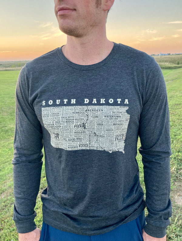 A man wearing a unisex, long sleeved, Dark Grey Heather, cotton polyester tee shirt. The front graphic, done in light grey is of the state of South Dakota and most of its cities. Scratchpad Tees original design available in sizes small to 3X-Large.