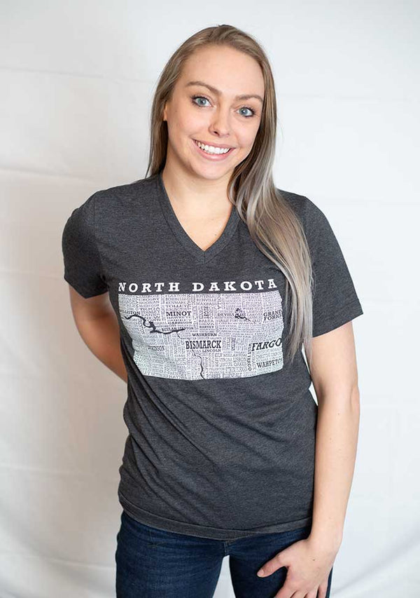 A woman wearing a unisex sized,Dark Grey Heather V neck, cotton polyester, short sleeve tee. Front white graphic is of North Dakota and its cities. Sizes small to 3X-Lagre. A Scratchpad Tees original design