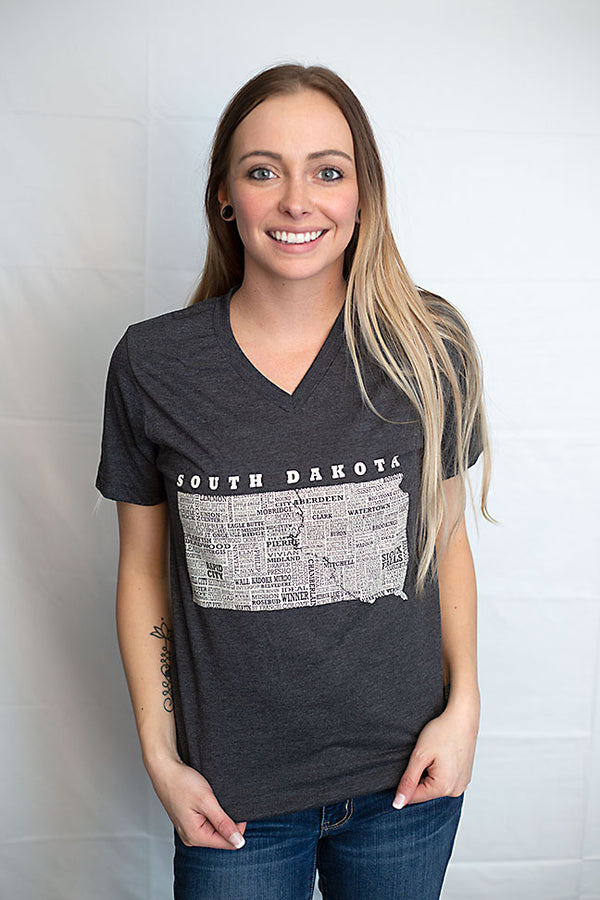 A woman wearing a unisex sized, Dark Grey Heather V neck, short sleeve tee. Front white graphic is of South Dakota and its cities. Sizes small to 3X-Lagre. A Scratchpad Tees original design
