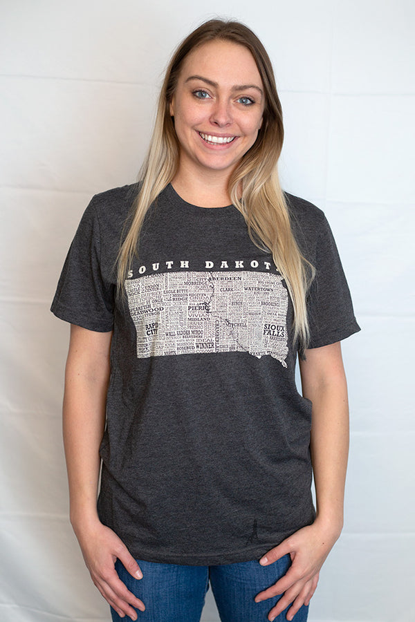 A woman wearing a unisex sized,Dark Grey Heather crew neck, cotton polyester, short sleeve tee. Front white graphic is of South Dakota and its cities. Sizes small to 3X-Large. A Scratchpad Tees original design.