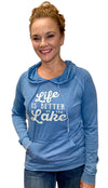 A woman in a featherweight, pouch front, hooded long sleeved sweatshirt. This Denim Blue shirt has a light grey graphic done in light grey with a playful font that reads Life Is Better At The Lake. 
