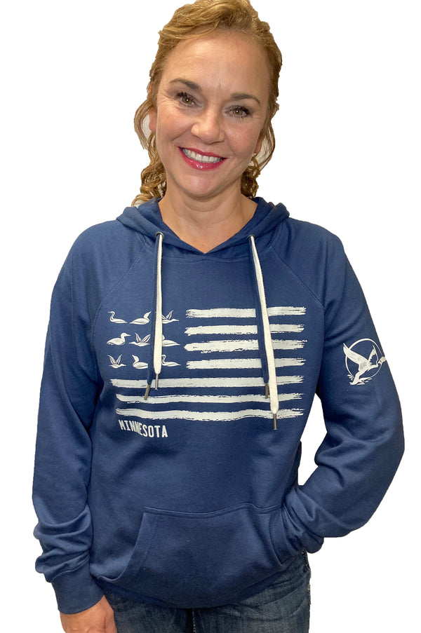 A woman wearing an Indigo colored, hooded, pouch front sweatshirt. Front graphic in light grey shows 9 loons and a field of stripes (looks like a flag). Minnesota is printed under the stripes field. There is a loon design feature on the left sleeve.