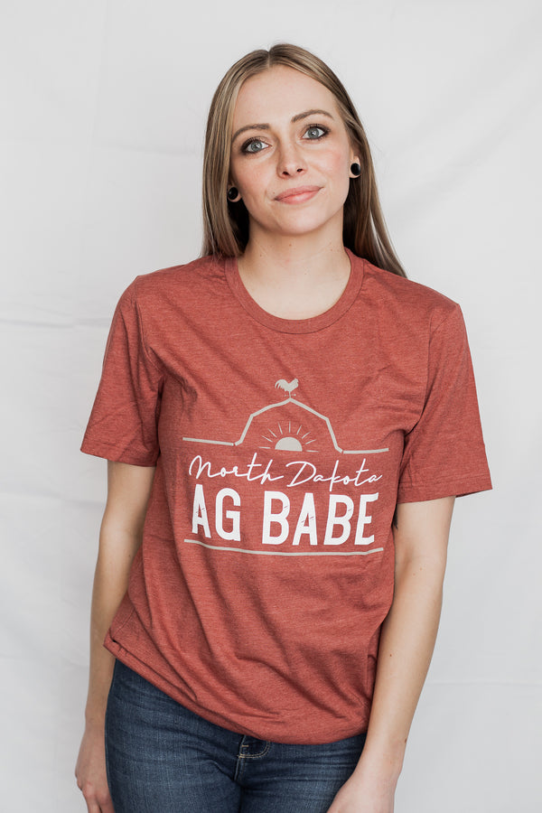 A short sleeved unisex crew neck tee shirt in Heather Clay color. The graphic shows a rooster perched on a barn roof at sunrise. The words North Dakota Ag Babe are printed on the shirt.