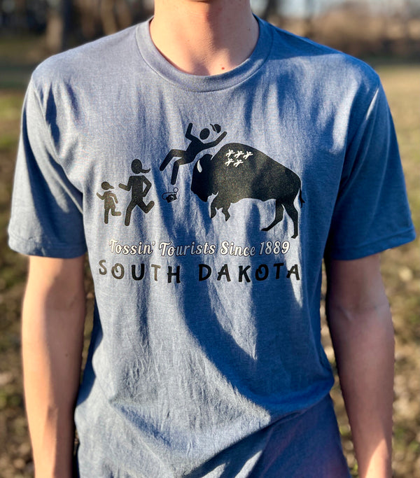 A male model wearing a Flint Blue Trim-blend crew neck tee shirt. The front graphic includes a charging buffalo launching a tourist family up in the air. The caption reads; Tossin' Tourists since 1889 in South Dakota.