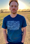 A male model wearing a Steel Blue, unisex, 100 percent cotton, crew neck short sleeved tee. The white front graphic is a map of South Dakota counties with historical number and name. Wording reads South Dakota, The Mount Rushmore State. Available in sizes Small to 3 X-Large