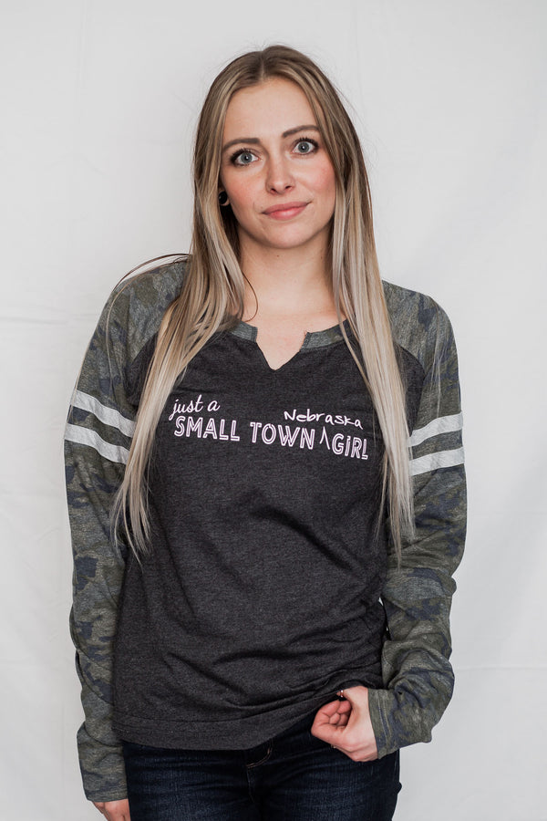 A ladies cut long sleeve notched collar jersey styled tee shirt. Dark grey body with Camo sleeves . Front white graphic says Just A Small Town Nebraska Girl .