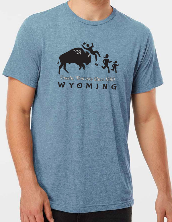 A male model wearing a Flint Blue Trim-blend crew neck tee shirt. The front graphic includes a charging buffalo launching a tourist family up in the air. The caption reads; Tossin' Tourists since 1890 in Wyoming.